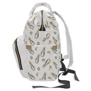 May include: A white backpack with a repeating pattern of Peter Rabbit illustrations, carrots, and cabbages. It has a white handle, straps, and a side pocket with gold-coloured trim. The zip is also gold-coloured.