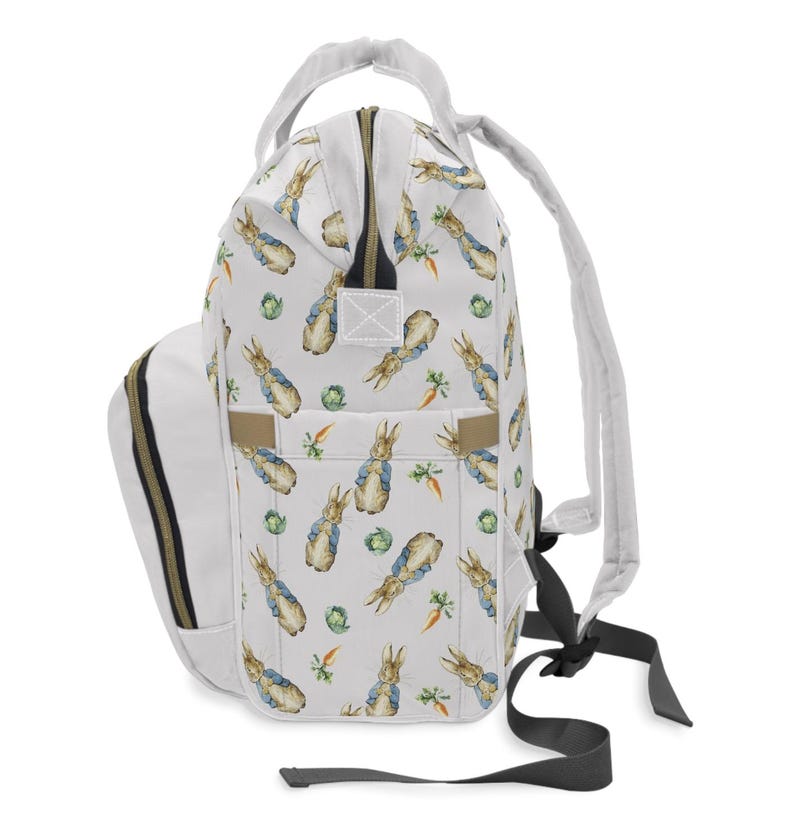 May include: A white backpack with a repeating pattern of Peter Rabbit illustrations, carrots, and cabbages. It has a white handle, straps, and a side pocket with gold-coloured trim. The zip is also gold-coloured.