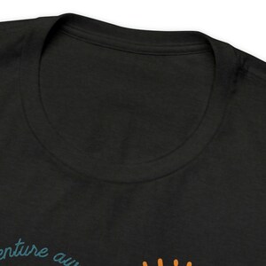May include: Black t-shirt with a crew neck. The shirt features the text "adventure awaits" in teal script, with an orange graphic below. The shirt is made of a soft material.