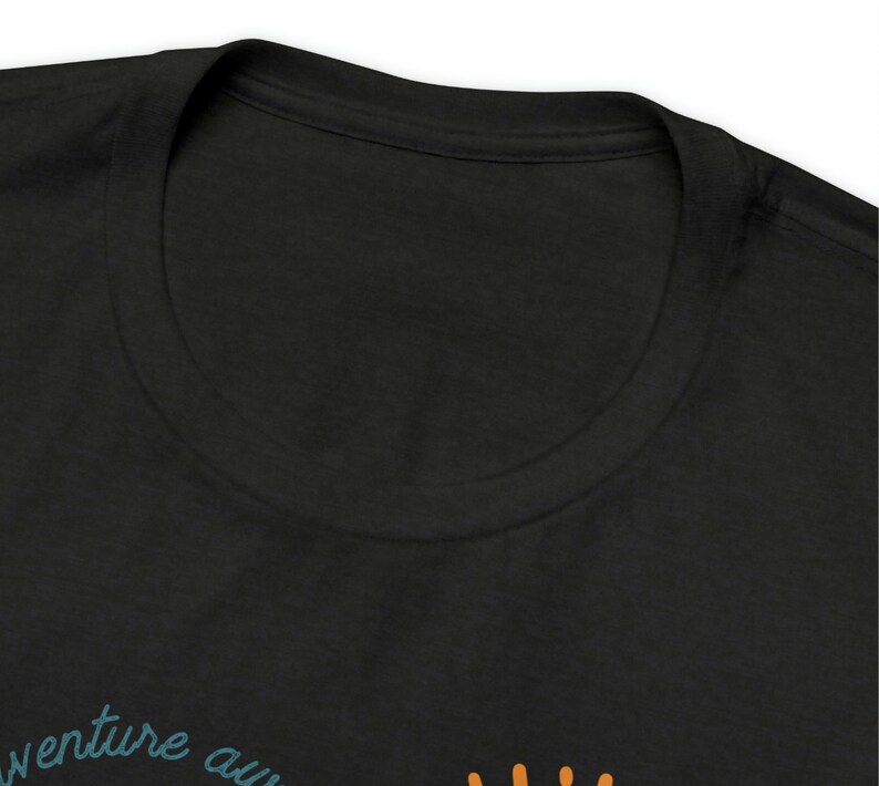 May include: Black t-shirt with a crew neck. The shirt features the text "adventure awaits" in teal script, with an orange graphic below. The shirt is made of a soft material.