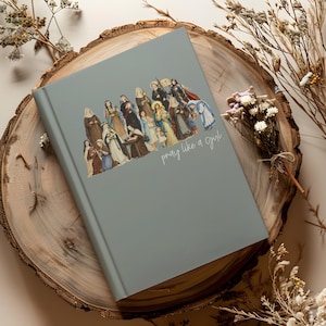 May include: A gray journal with an illustration of various religious figures. The cover features the phrase "pray like a girl" in cursive script. The journal is resting on a wooden slice, with dried flowers in the background.