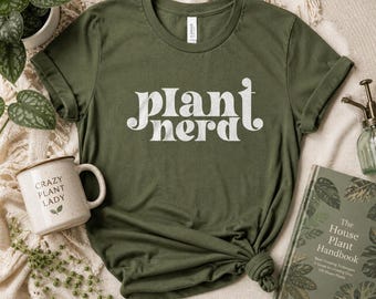 Puede incluir: Camiseta verde oliva con las palabras "Plant Nerd" en blanco, con una fuente estilizada. Una taza blanca con las palabras "Crazy Plant Lady" está al lado de la camiseta. También se ve un libro titulado "The House Plant Handbook".