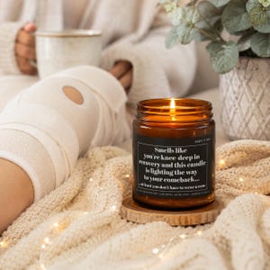 Op de afbeelding: Een bruine glazen kaars met een zwart etiket met de witte tekst: Smells like you're knee-deep in recovery and this candle is lighting the way to your comeback... at least you don't have to wear a cone. De kaars staat op een houten schijf.