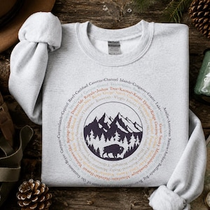 May include: A light gray crewneck sweatshirt featuring a circular graphic design. The center displays a dark purple illustration of mountains, pine trees, and a bison. Surrounding the graphic are the names of various national parks in a circle.
