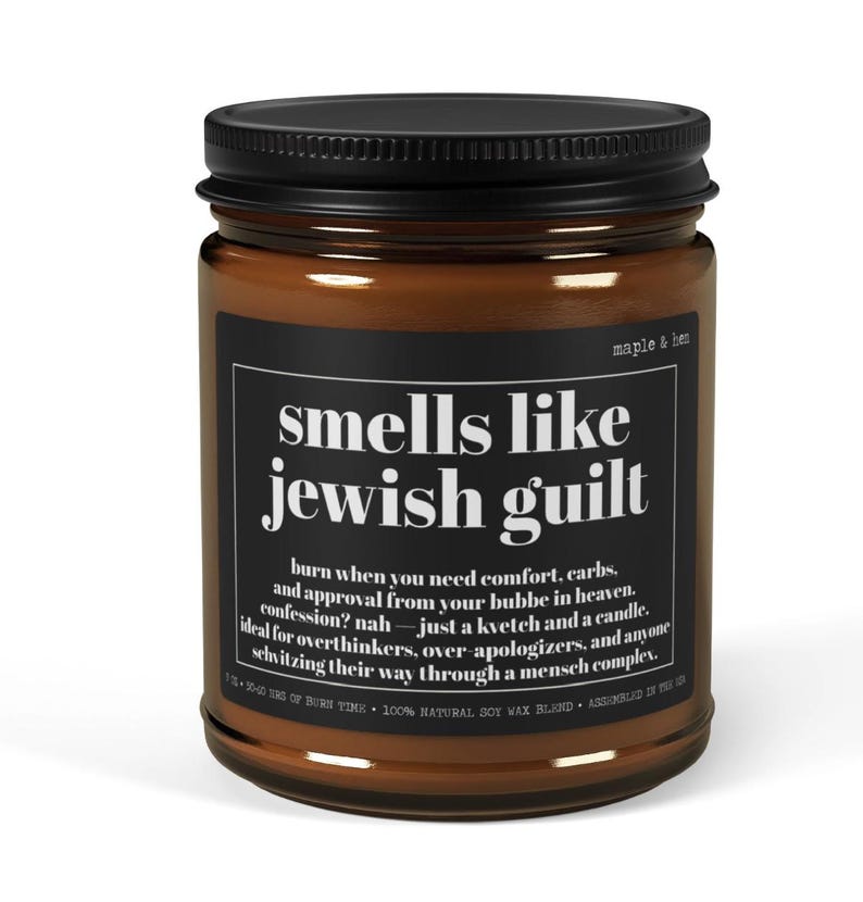 May include: A brown glass candle jar with a black lid and a black label featuring white text that reads: smells like jewish guilt, burn when you need comfort, carbs, and approval from your bubbe in heaven. confession? nah &mdash; just a kvetch and a candle.