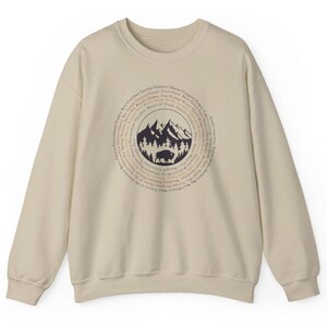 May include: A beige crewneck sweatshirt featuring a dark brown circular graphic on the chest. The design includes a central illustration of mountains, a forest, and a bison, surrounded by a ring of text listing various national park names.