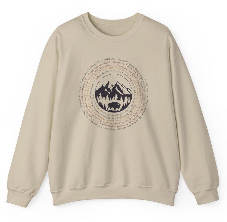 May include: A beige crewneck sweatshirt featuring a dark brown circular graphic on the chest. The design includes a central illustration of mountains, a forest, and a bison, surrounded by a ring of text listing various national park names.