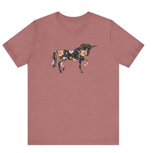 Puede incluir: Una camiseta de color rosa polvoriento con un dise&ntilde;o floral de unicornio. La silueta del unicornio est&aacute; llena de un patr&oacute;n de flores rosas, amarillas y blancas sobre un fondo verde oscuro. La camiseta tiene mangas cortas y cuello redondo.