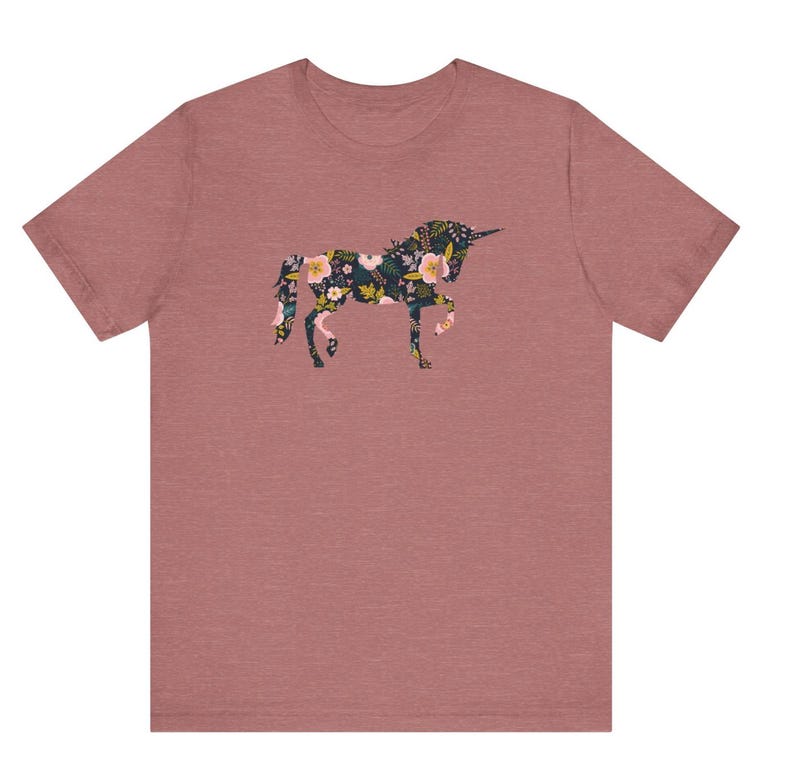 Puede incluir: Una camiseta de color rosa polvoriento con un dise&ntilde;o floral de unicornio. La silueta del unicornio est&aacute; llena de un patr&oacute;n de flores rosas, amarillas y blancas sobre un fondo verde oscuro. La camiseta tiene mangas cortas y cuello redondo.