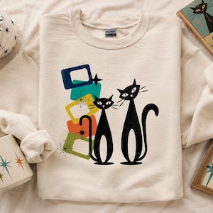 May include: Cream-colored sweatshirt featuring a retro design with two black cat silhouettes and colorful geometric shapes. The design includes blue, green, orange, and yellow elements, evoking a mid-century modern aesthetic. A matching mug and book are also visible.