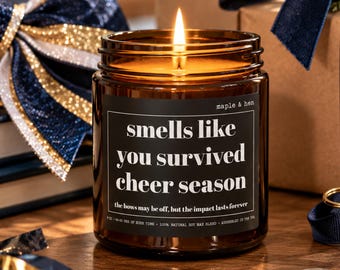 May include: A lit candle in an amber glass jar with a black label featuring the white text: smells like you survived cheer season, the bows may be off, but the impact lasts forever, maple & hen, 9 oz, 30-40 hrs of burn time, 100% natural soy wax blend.