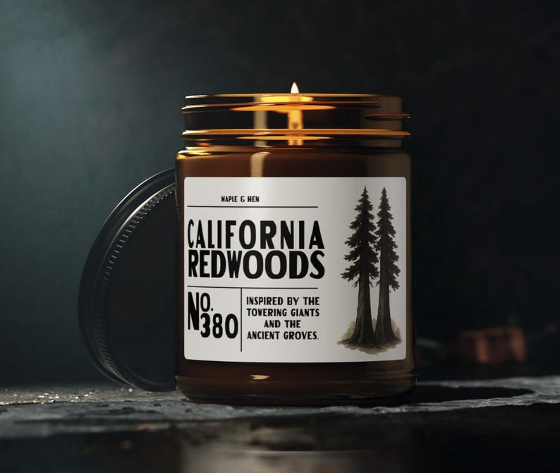 May include: A lit candle in a brown glass jar with a gold lid. The label reads "CALIFORNIA REDWOODS" with the text "INSPIRED BY THE TOVERING GIANTS AND THE ANCIENT GROVES." The jar is next to a black lid.