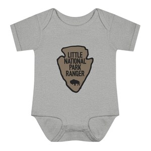 May include: A heather grey short-sleeve baby bodysuit featuring a tan arrowhead-shaped graphic on the chest. The graphic contains the black text LITTLE NATIONAL PARK RANGER above a small black silhouette of a bison, all outlined in black.
