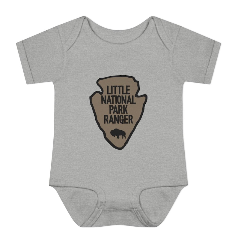 May include: A heather grey short-sleeve baby bodysuit featuring a tan arrowhead-shaped graphic on the chest. The graphic contains the black text LITTLE NATIONAL PARK RANGER above a small black silhouette of a bison, all outlined in black.