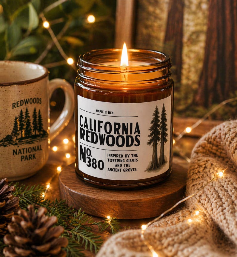 May include: A lit candle in a glass jar with a label that reads "California Redwoods." The candle sits on a wooden stand, next to a mug that says "Redwoods National Park." The scene is decorated with pinecones and fairy lights.