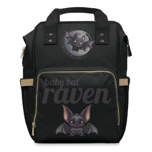 May include: Black diaper bag with a cartoon bat design. The bag features a large front pocket with the words "baby bat raven" printed in a stylized font. The bag has a top handle, side pockets, and gold-colored zippers.