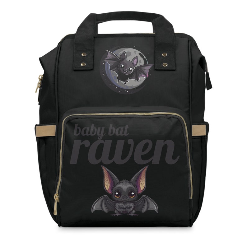 May include: Black diaper bag with a cartoon bat design. The bag features a large front pocket with the words "baby bat raven" printed in a stylized font. The bag has a top handle, side pockets, and gold-colored zippers.