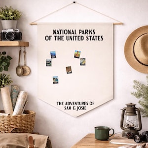 May include: A cream-colored fabric wall banner featuring the black text NATIONAL PARKS OF THE UNITED STATES and THE ADVENTURES OF SAM & JOSIE. The banner is suspended from a wooden rod with a string and displays several small rectangular park pins.