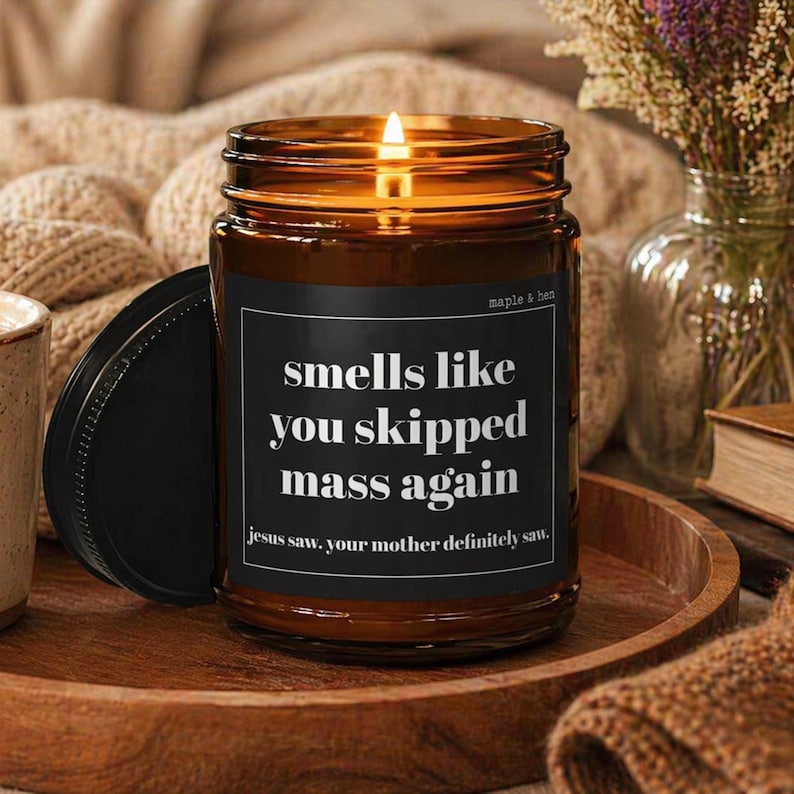 May include: A lit brown glass jar candle with a black lid. The candle has a black label with white text that reads "smells like you skipped mass again." The text "jesus saw. your mother definitely saw." is also on the label. The candle is on a wooden tray.