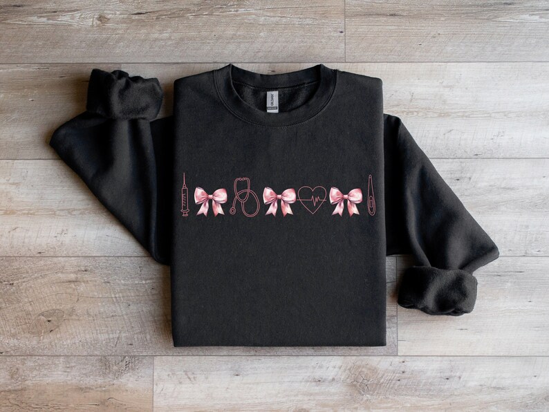 May include: Black sweatshirt with a pink medical-themed design. The design includes a syringe, bows, a stethoscope, a heart with a heartbeat line, and a thermometer. The sweatshirt is made of a soft material.