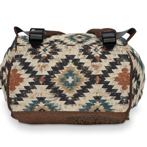 May include: A duffel bag with a Southwestern geometric pattern in cream, black, teal, and rust. The bag has brown leather accents on the bottom and straps, with black plastic buckles. The bag is designed for travel or carrying gear.