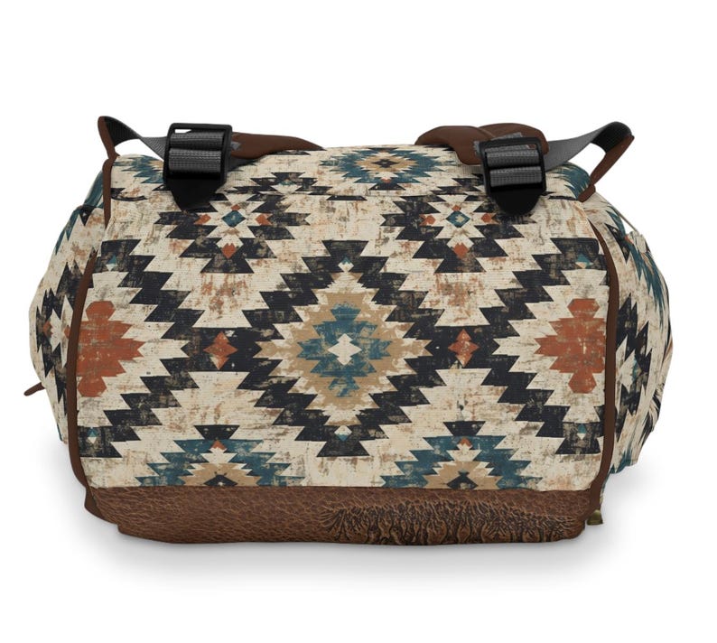 May include: A duffel bag with a Southwestern geometric pattern in cream, black, teal, and rust. The bag has brown leather accents on the bottom and straps, with black plastic buckles. The bag is designed for travel or carrying gear.
