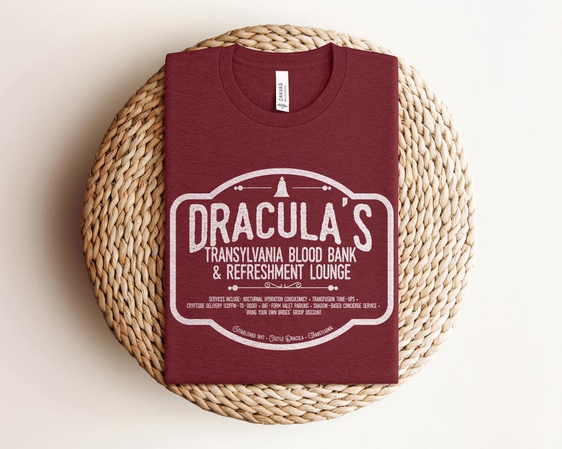May include: A maroon t-shirt with a white graphic that reads "DRACULA'S TRANSYLVANIA BLOOD BANK & REFRESHMENT LOUNGE". The shirt is folded on a round, woven, beige placemat. The shirt has a crew neck and short sleeves.
