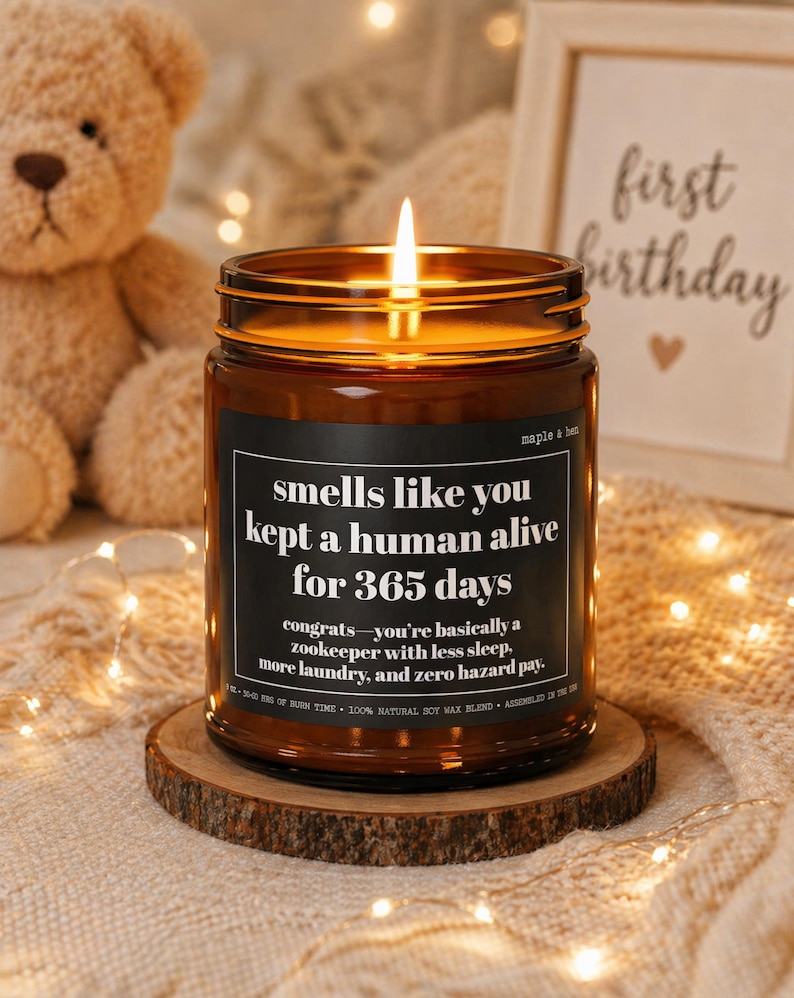 May include: A lit candle in a brown glass jar with a black label that reads "smells like you kept a human alive for 365 days." The candle sits on a wooden coaster. A teddy bear and a framed picture are in the background.