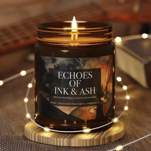 May include: A lit brown glass jar candle with the text "ECHOES OF INK & ASH". The candle sits on a wooden coaster, surrounded by string lights. The candle's label includes the phrases "SMOKE | INK-STAINED PARCHMENT | EMBER GLOW".