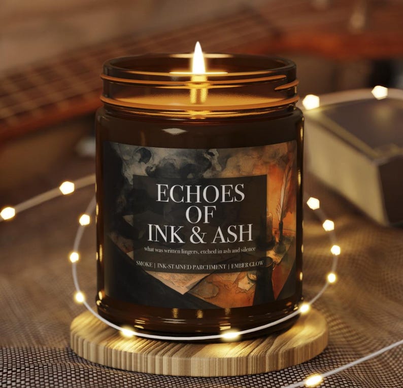 May include: A lit brown glass jar candle with the text "ECHOES OF INK & ASH". The candle sits on a wooden coaster, surrounded by string lights. The candle's label includes the phrases "SMOKE | INK-STAINED PARCHMENT | EMBER GLOW".