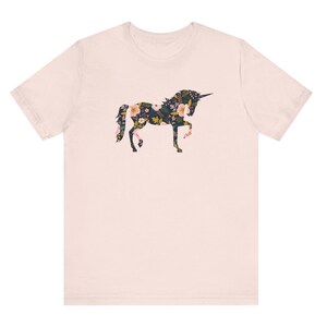 Puede incluir: Una camiseta rosa claro con un dise&ntilde;o de unicornio floral. La silueta del unicornio est&aacute; llena de un patr&oacute;n de flores rosas, amarillas y blancas sobre un fondo oscuro. La camiseta tiene mangas cortas y cuello redondo.