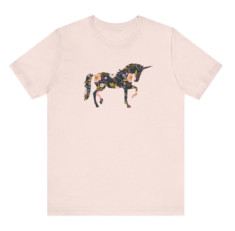 Puede incluir: Una camiseta rosa claro con un dise&ntilde;o de unicornio floral. La silueta del unicornio est&aacute; llena de un patr&oacute;n de flores rosas, amarillas y blancas sobre un fondo oscuro. La camiseta tiene mangas cortas y cuello redondo.
