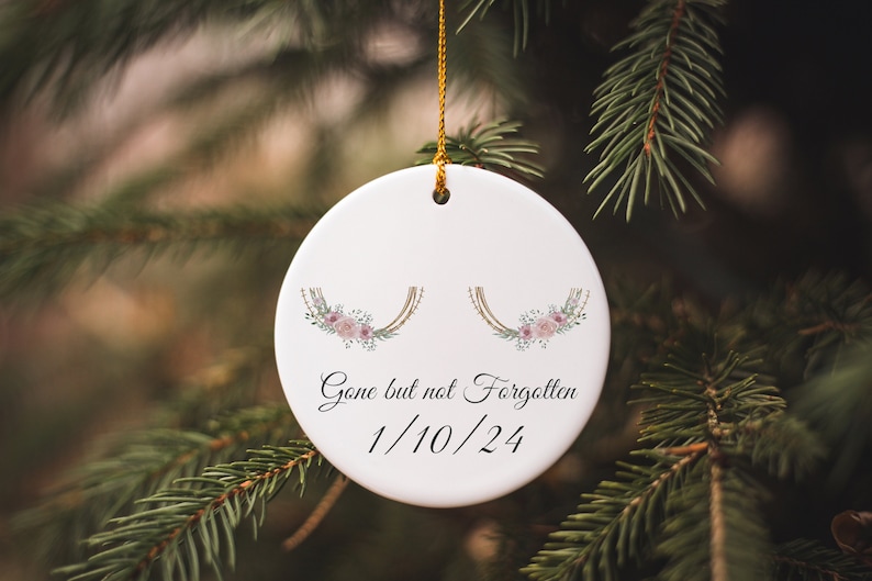 May include: White ceramic ornament with a gold string, featuring a floral design with pink flowers and green leaves. The ornament has the text "Gone but not Forgotten 1/10/24" written on it.