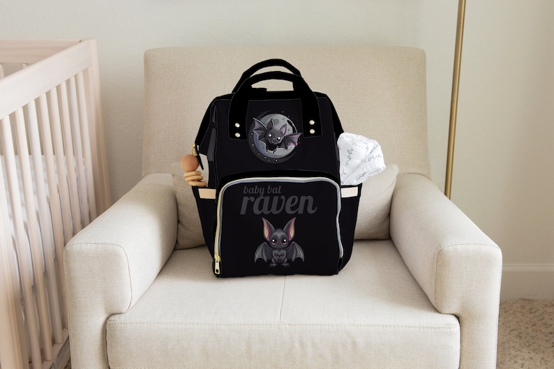 May include: Black diaper bag with a bat theme. The bag features a cartoon bat illustration and the text "baby bat raven". It has multiple pockets, a top handle, and a zipper closure. A white cloth and a wooden toy are visible.