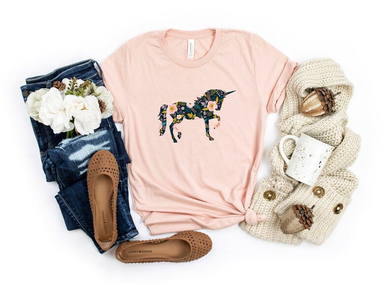 Puede incluir: Una camiseta rosa claro con un dise&ntilde;o floral de unicornio. La camiseta se combina con vaqueros azules, bailarinas marrones, una bufanda color crema y una taza blanca. El dise&ntilde;o del unicornio es una silueta llena de flores coloridas.