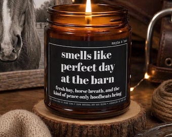 May include: A lit candle in an amber glass jar with a black label that reads: smells like perfect day at the barn, fresh hay, horse breath, and the kind of peace only hoofbeats bring. The candle sits on a circular wooden slice base.