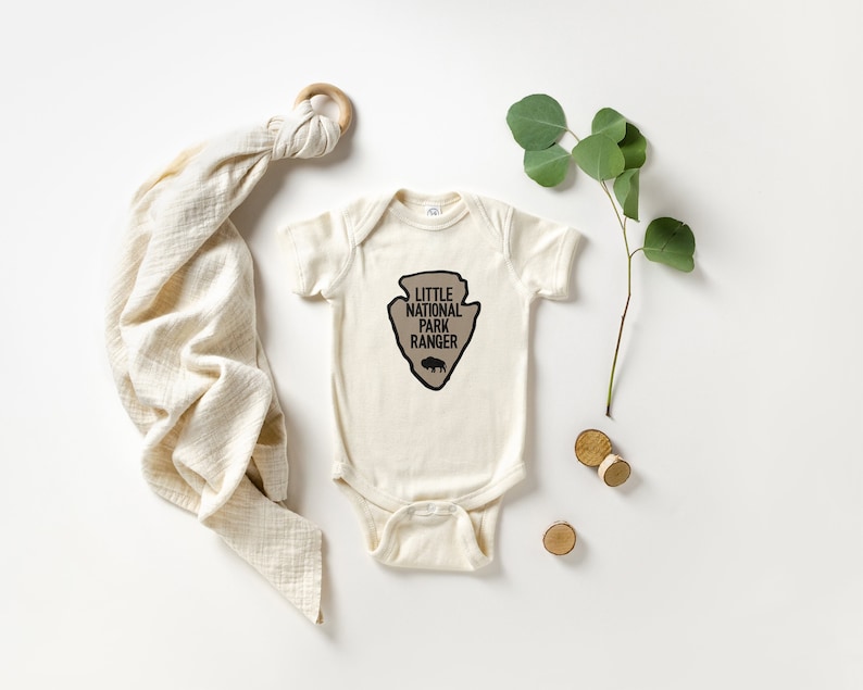 May include: An off-white baby bodysuit featuring a tan arrowhead-shaped graphic with the text "LITTLE NATIONAL PARK RANGER" and a small bison silhouette printed in black. The garment has short sleeves and a snap-button closure at the bottom.