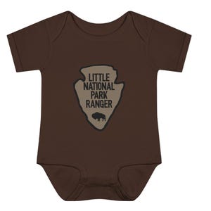 May include: A dark brown short-sleeved baby bodysuit featuring a tan arrowhead-shaped graphic on the chest. The graphic contains the black text "LITTLE NATIONAL PARK RANGER" stacked above a small black silhouette of a standing bison.