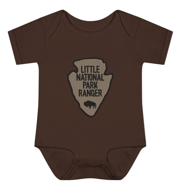 May include: A dark brown short-sleeved baby bodysuit featuring a tan arrowhead-shaped graphic on the chest. The graphic contains the black text "LITTLE NATIONAL PARK RANGER" stacked above a small black silhouette of a standing bison.