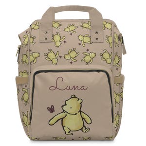 May include: A beige backpack featuring a repeating pattern of yellow cartoon bears. The front pocket displays a larger bear illustration with the name "Luna" in cursive. The backpack has side pockets and a top handle.