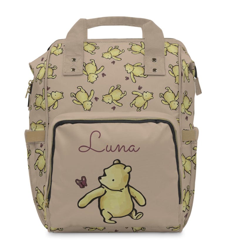 May include: A beige backpack featuring a repeating pattern of yellow cartoon bears. The front pocket displays a larger bear illustration with the name "Luna" in cursive. The backpack has side pockets and a top handle.