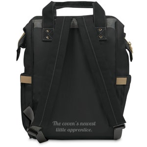 May include: Black backpack with gray and tan accents. Features adjustable shoulder straps, a top handle, and side pockets. The text "The coven's newest little apprentice" is printed on the back.