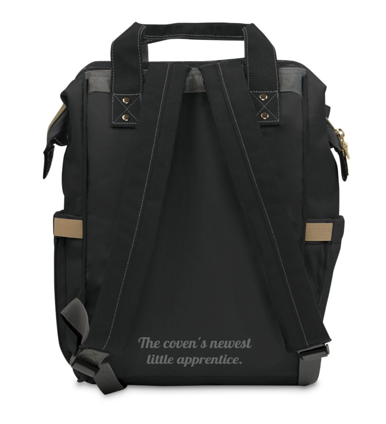 May include: Black backpack with gray and tan accents. Features adjustable shoulder straps, a top handle, and side pockets. The text "The coven's newest little apprentice" is printed on the back.