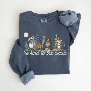 May include: A slate blue crewneck sweatshirt featuring a graphic of a raccoon, a bear, and a fox in a forest setting with a campfire and moon. The text "be kind to the locals" is printed in white script below the illustrated woodland animals.
