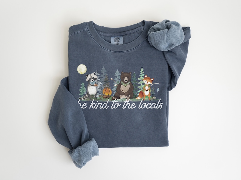 May include: A slate blue crewneck sweatshirt featuring a graphic of a raccoon, a bear, and a fox in a forest setting with a campfire and moon. The text "be kind to the locals" is printed in white script below the illustrated woodland animals.