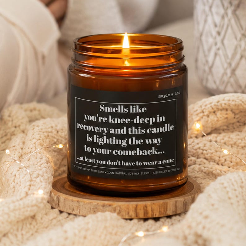 Op de afbeelding: Een brandende kaars in een amberkleurige glazen pot met een zwart etiket met de witte tekst: Smells like you're knee-deep in recovery and this candle is lighting the way to your comeback... at least you don't have to wear a cone.