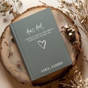 May include: A gray journal with the text "dear dad," and "A JOURNAL FOR ALL THE THINGS I STILL WANT TO TELL YOU" in white script. A white heart and the words "LOVE, SAMMY" are also on the cover. The journal is on a wooden surface.
