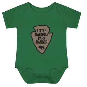 May include: A dark green short-sleeved baby bodysuit featuring a tan arrowhead-shaped graphic on the chest. The graphic contains the black text "LITTLE NATIONAL PARK RANGER" positioned above a small black silhouette of a standing bison.