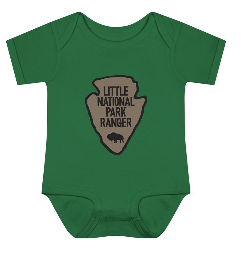 May include: A dark green short-sleeved baby bodysuit featuring a tan arrowhead-shaped graphic on the chest. The graphic contains the black text "LITTLE NATIONAL PARK RANGER" positioned above a small black silhouette of a standing bison.