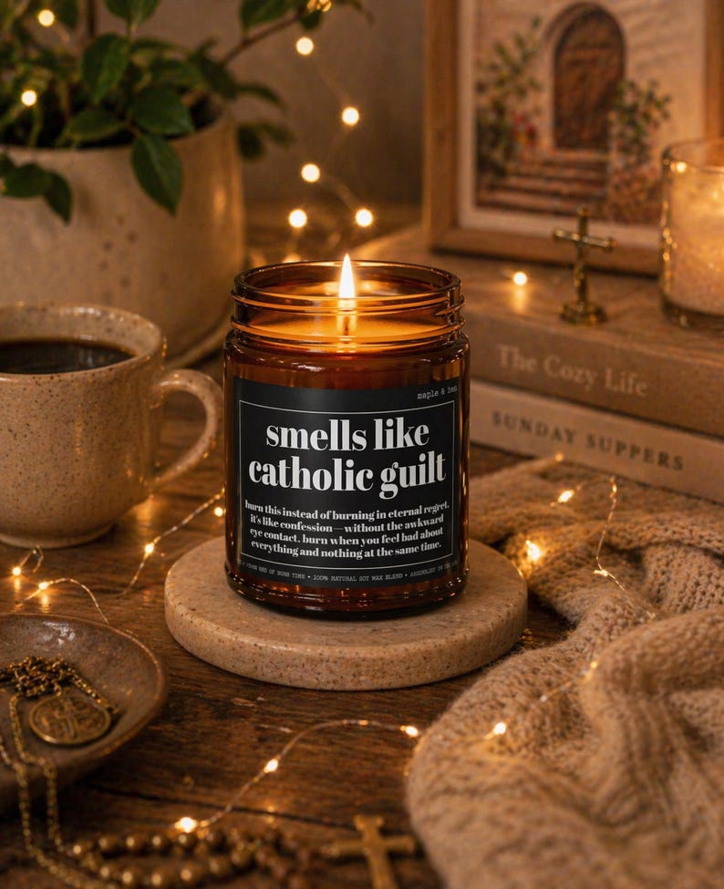 May include: A lit brown glass candle jar with the text "smells like catholic guilt" on a black label. The candle sits on a round stone coaster, with a cup of coffee and a decorative bowl in the background. The candle is surrounded by fairy lights.